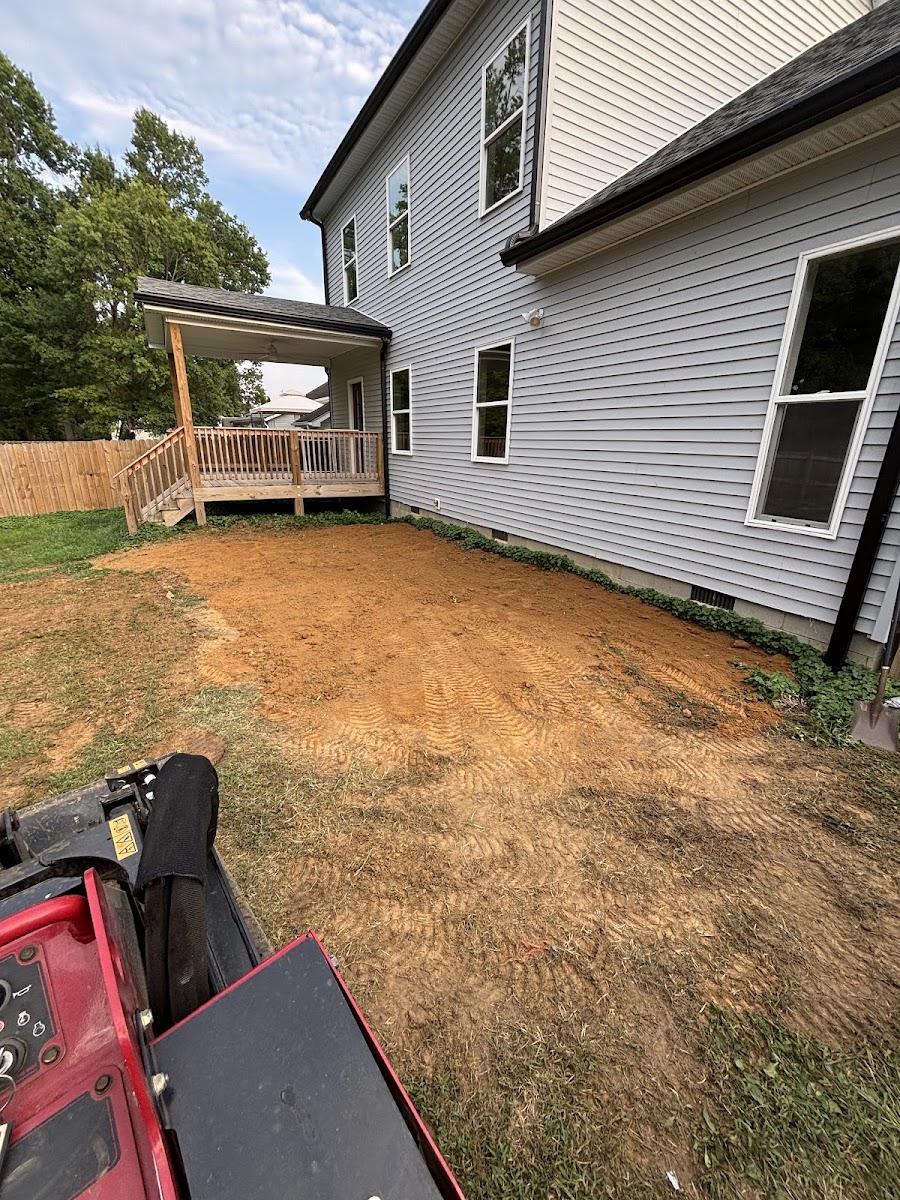 View from inside a compact track loader looking out over a freshly graded dirt yard behind a house.